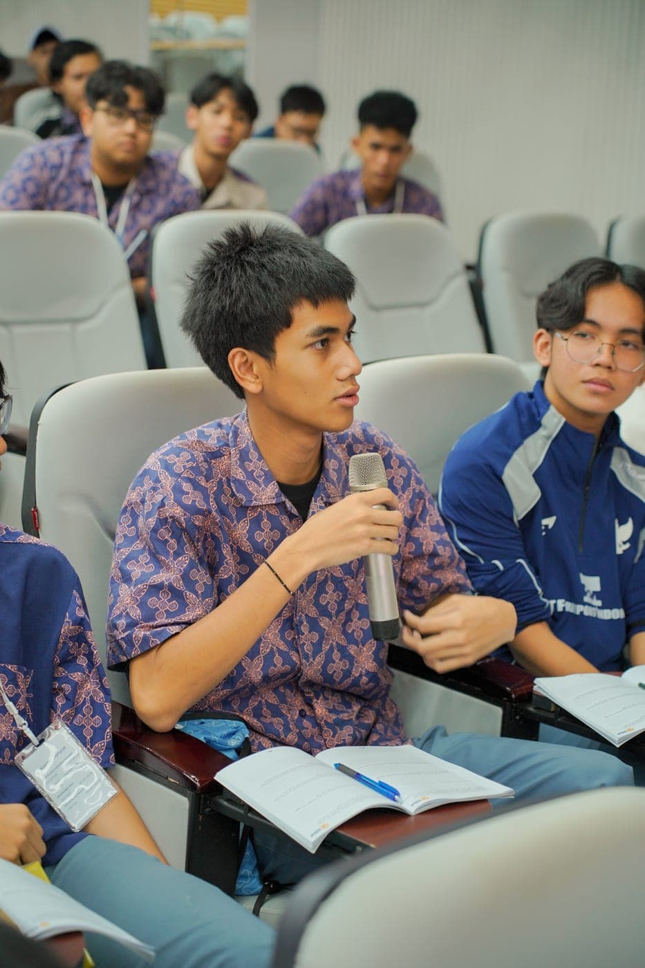 Seorang pelajar berdiri memegang mikrofon dan berbicara di tengah ruangan berisi peserta lain