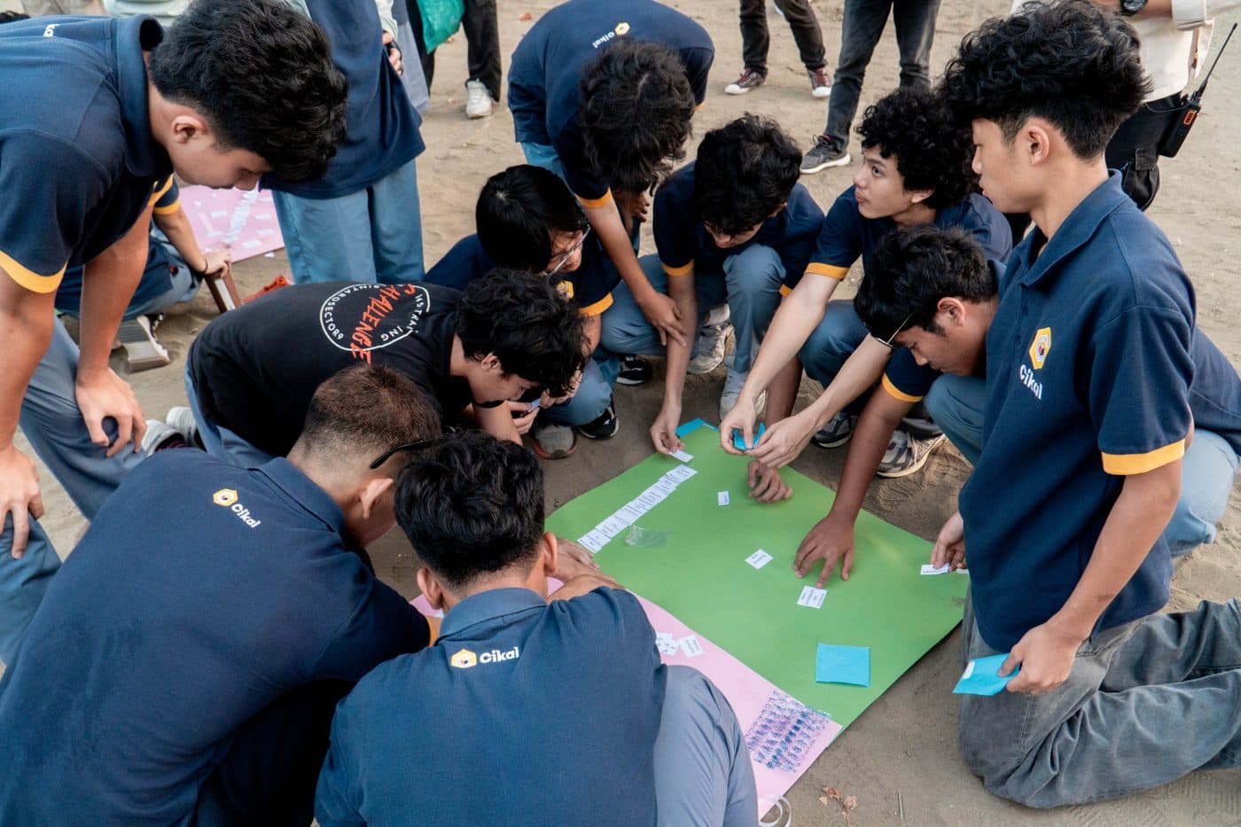 Sekelompok pelajar berkumpul melingkar sambil menunduk membahas strategi sebelum memulai tantangan di area terbuka